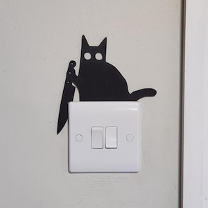 May include: A black cat silhouette holding a knife, positioned above a white light switch with two switches. The cat has large, round eyes and a tail. The wall is painted a light grey colour.