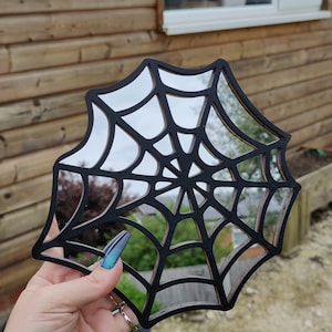 May include: A black wooden spiderweb mirror with a mirrored surface. The mirror is shaped like a spiderweb and has a black frame.