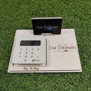 May include: A white wooden point-of-sale display featuring a SumUp card reader and a business card holder. The display is branded with "Lisa Engraves" and "Tap To Pay". A black business card holder reads "Take One".