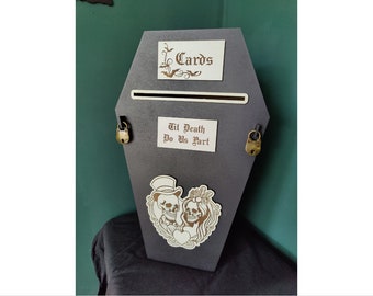 Può includere: Una scatola per biglietti di auguri nera a forma di bara con una fessura per i biglietti. La scatola ha un'etichetta bianca che dice "Cards" e un'altra etichetta bianca che dice "Til Death Do Us Part". La scatola ha due lucchetti dorati sui lati e una grafica di una coppia di scheletri sul davanti.