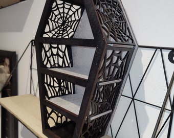 Può includere: Un ripiano in legno nero a forma di bara con tre ripiani e un design a ragnatela. Il ripiano è in legno e ha una finitura nera. I ripiani sono in materiale grigio chiaro. Il design a ragnatela è tagliato nel legno ed è visibile su tutti i lati del ripiano.