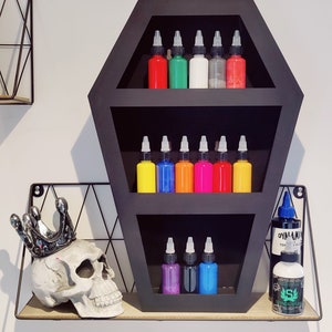 May include: A black coffin-shaped shelf with three shelves, filled with bottles of tattoo ink in various colours, including red, green, yellow, orange, pink, blue, and purple. The shelf is on a wooden surface with a skull wearing a crown on the left side and a bottle of green ink on the right side.