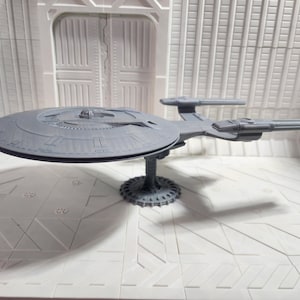 May include: A gray model of a spaceship with a circular main body and two long, rectangular extensions. The model is mounted on a detailed, circular base. The background is a white, geometric interior.