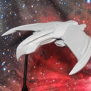 May include: A light gray model of a spacecraft, featuring a sleek, angular design with extended wing-like structures. The model is mounted on a black stand, set against a backdrop of a nebula and stars.