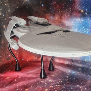 May include: A gray 3D printed model of a starship featuring a large circular saucer section, two rear engine nacelles, and a central hull structure. The model is supported by three thin black flexible stands that elevate it above the surface.