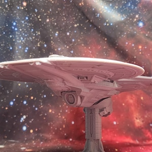 May include: A light gray, unpainted 3D printed model of a starship featuring a large circular primary hull, a central deflector dish, and two rear engine nacelles supported by a vertical stand, showcasing intricate paneling and structural details.