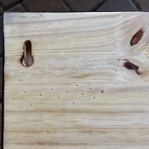 May include: A close-up of a light wood sign with a small metal hook on the back. The wood has a natural grain and a few knots. There are also several small holes in the wood, likely from previous use.