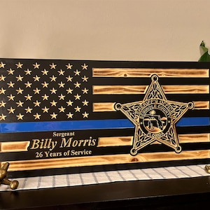 May include: A wooden American flag with a blue line, a sheriff's star, and the text "Sergeant Billy Morris 26 Years of Service".