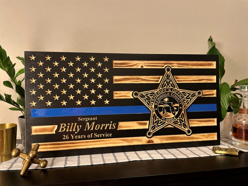 May include: A wooden American flag with a blue line, a sheriff's star, and the text "Sergeant Billy Morris 26 Years of Service".