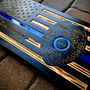 May include: A wooden American flag with a blue line and a police badge. The badge has the text "DETECTIVE FORT MYERS POLICE FL" and the number "6015". The name "D. Rosenberger" is engraved on the flag.