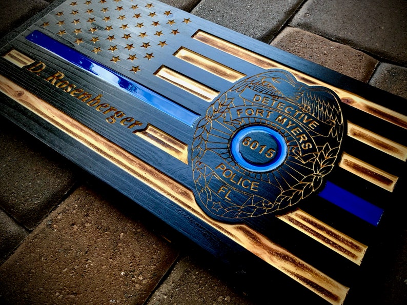 May include: A wooden American flag with a blue line and a police badge. The badge has the text "DETECTIVE FORT MYERS POLICE FL" and the number "6015". The name "D. Rosenberger" is engraved on the flag.