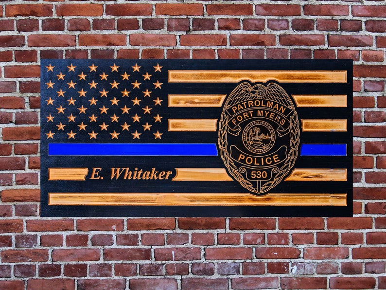 May include: Wooden American flag with a blue line, a police badge with the text "Patrolman Fort Myers Police 530" and the name "E. Whitaker" engraved on the bottom.