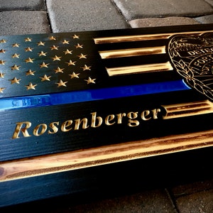 May include: A wooden sign with a black and gold American flag design, a blue line, and a police badge. The sign is personalized with the name "D. Rosenberger".