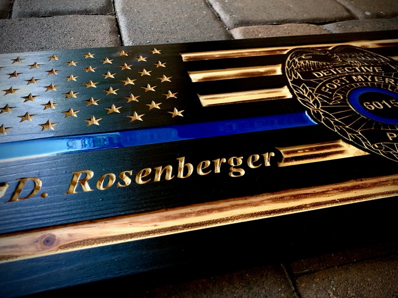 May include: A wooden sign with a black and gold American flag design, a blue line, and a police badge. The sign is personalized with the name "D. Rosenberger".