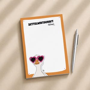 Könnte beinhalten: Ein rechteckiger Notizblock mit orangefarbenem Rand und weißer Mitte, der oben den Text ZETTELWIRTSCHAFT deluxe und unten eine weiße Gans mit einer rosa herzförmigen Sonnenbrille zeigt, begleitet von einem silbernen Kugelschreiber.