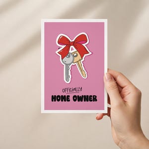Könnte beinhalten: Eine rechteckige Grußkarte mit rosa Hintergrund und einer grafischen Illustration von zwei Metallschlüsseln, die mit einer roten Schleife zusammengebunden sind. Unter den Schlüsseln steht der Text "OFFICIALLY HOME OWNER" in schwarzer, fetter Schrift.