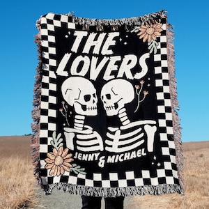 Può includere: Coperta a scacchi neri e bianchi con una grafica di due scheletri che si abbracciano. Gli scheletri sono etichettati "Jenny & Michael" e il testo "The Lovers" è sopra di loro.