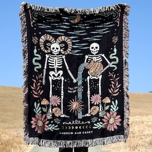 May include: Black woven tapestry with two skeletons holding hands. The skeletons are wearing floral crowns and the tapestry has a floral border. The text "Till death do us part" is written in the center of the tapestry. The tapestry also has the text "Andrew and Casey" at the bottom.