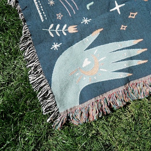 May include: A woven throw blanket with a dark blue background and a light blue hand with a sun and moon design. The blanket has a fringe border with multicolored threads.