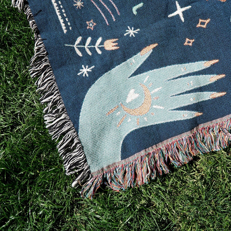 May include: A woven throw blanket with a dark blue background and a light blue hand with a sun and moon design. The blanket has a fringe border with multicolored threads.