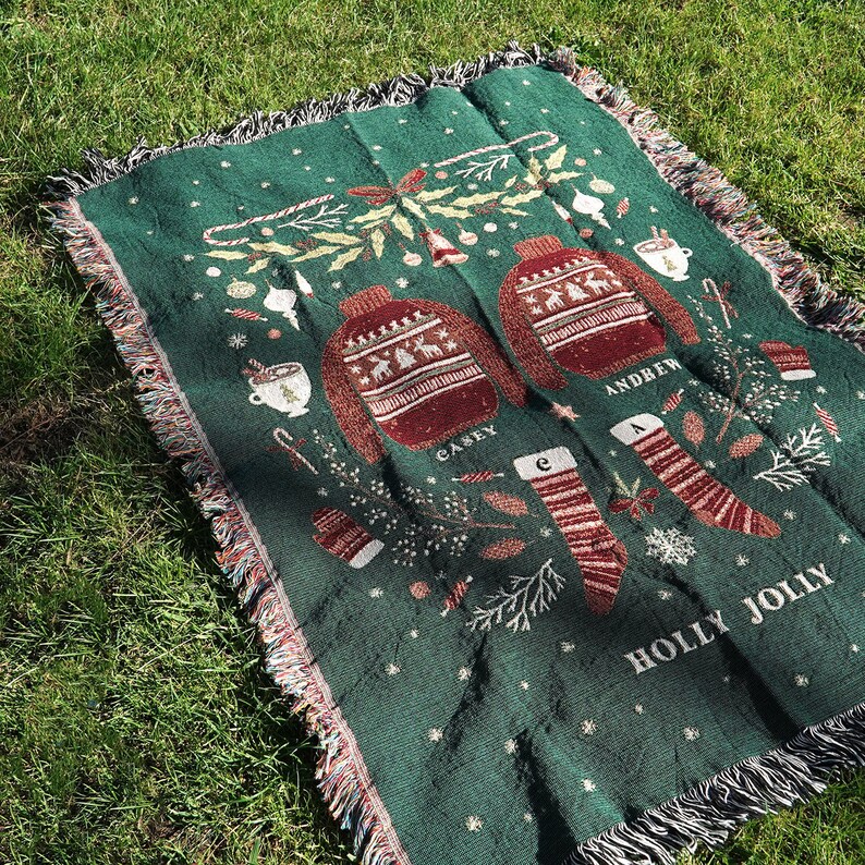 May include: Green throw blanket with a festive Christmas design featuring two red and white sweaters with the names "Casey" and "Andrew" embroidered on them. The blanket also features a holly sprig, candy canes, and the text "Holly Jolly".