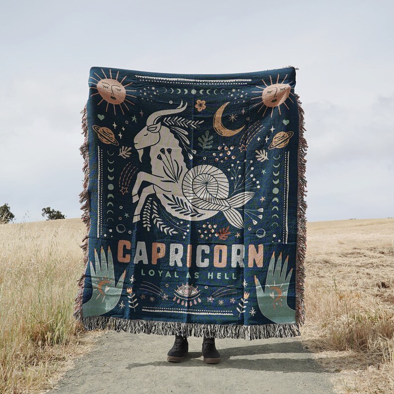 May include: A dark teal woven throw blanket featuring a Capricorn zodiac design. The design includes a goat, celestial elements, and the text "CAPRICORN LOYAL AS HELL". The blanket has fringed edges and is held up against a natural outdoor backdrop.