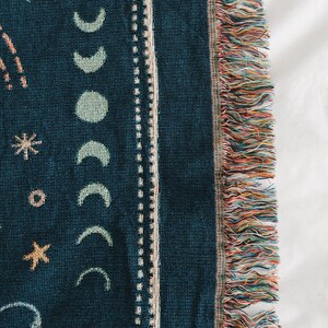 May include: Dark teal throw blanket with a celestial design. The blanket features crescent moons, stars, and other celestial shapes in light teal and beige. The edge has a fringe with multiple colors.