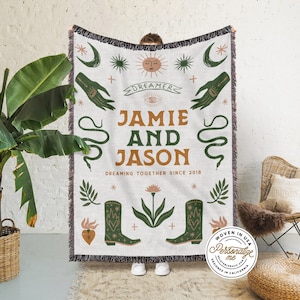 Può includere: Una coperta in tessuto intrecciato con sfondo bianco e accenti verdi, marroni e arancioni. La coperta presenta un design fantasioso con un sole, una luna, stelle, mani, un serpente, stivali e un fiore. Il testo "Dreamer", "Jamie e Jason", e "Dreaming Together Since 2018" è stampato sulla coperta.