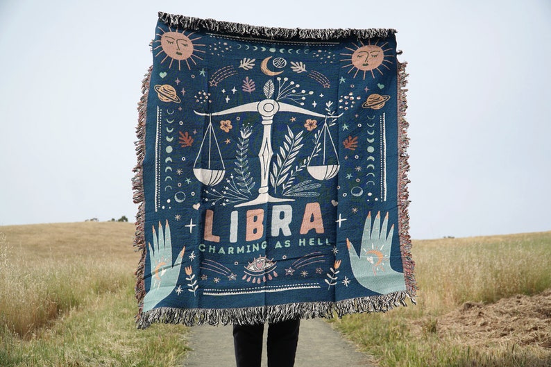 May include: A blue woven tapestry with a Libra zodiac sign and the text "Libra Charming as Hell". The tapestry features a scale, stars, moons, and floral designs.
