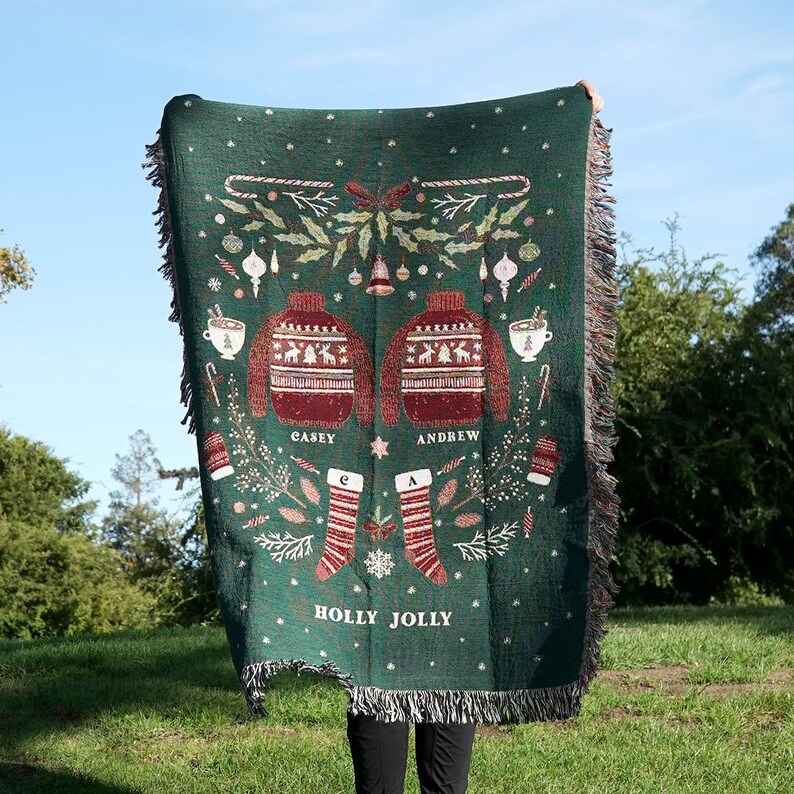 May include: A green woven throw blanket with a Christmas theme. It features two red and white sweaters with the names "Casey" and "Andrew" on them. The blanket also has a holly jolly design with stockings, candy canes, and other festive elements.