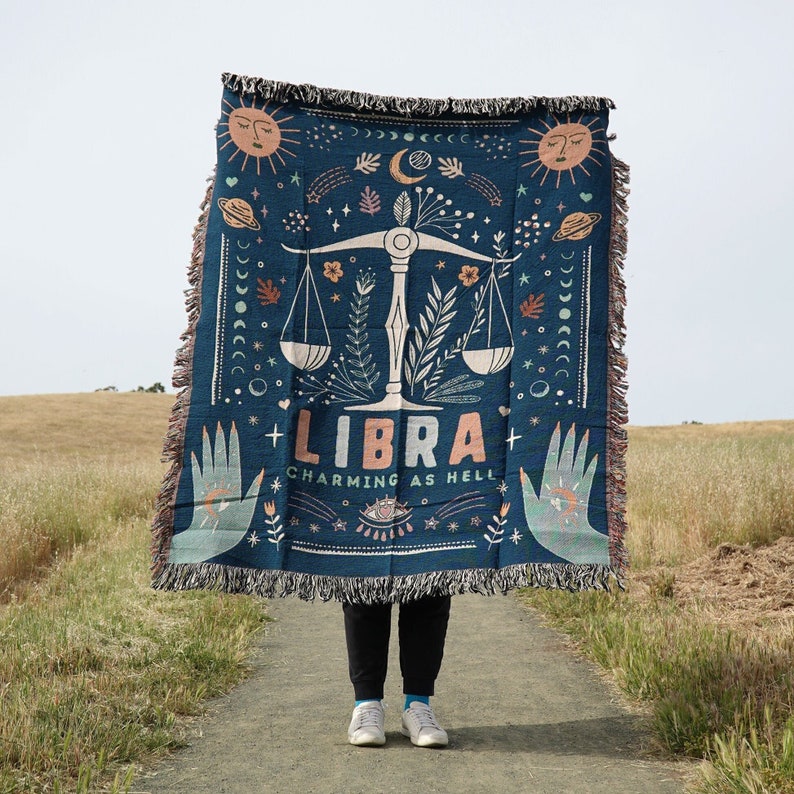 May include: A blue woven tapestry with a Libra zodiac symbol and the text "Libra Charming as Hell". The tapestry features a scale, stars, and moons.