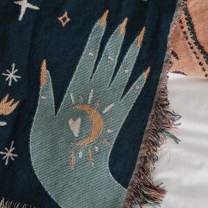 May include: A woven throw blanket with a teal hand design featuring gold fingernails, a crescent moon, and a sunburst. The blanket has a dark blue background with fringe along the edge. The design includes stars and other celestial elements.