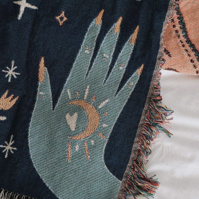 May include: A woven throw blanket with a teal hand design featuring gold fingernails, a crescent moon, and a sunburst. The blanket has a dark blue background with fringe along the edge. The design includes stars and other celestial elements.
