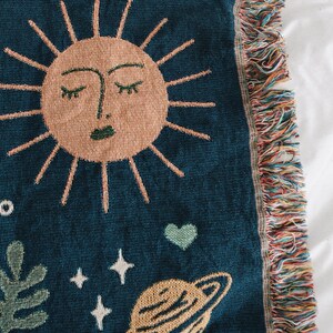May include: A woven tapestry with a blue background featuring a stylized sun with a closed eye design, a green heart, a planet with rings, and white stars.