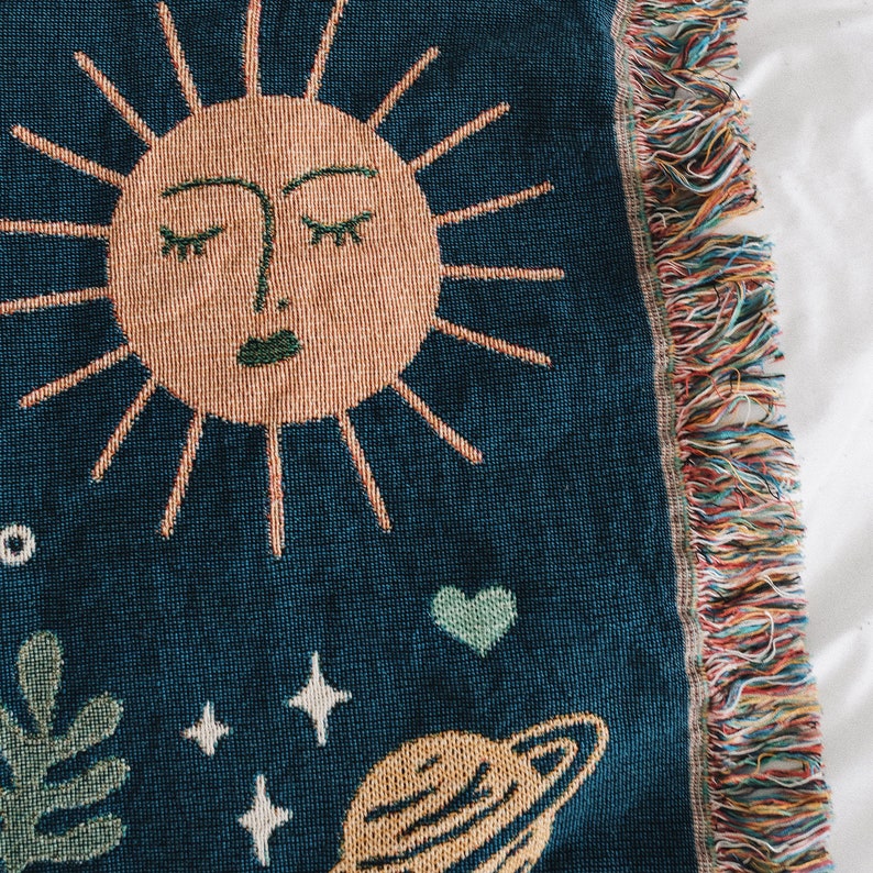 May include: A woven tapestry with a blue background featuring a stylized sun with a closed eye design, a green heart, a planet with rings, and white stars.