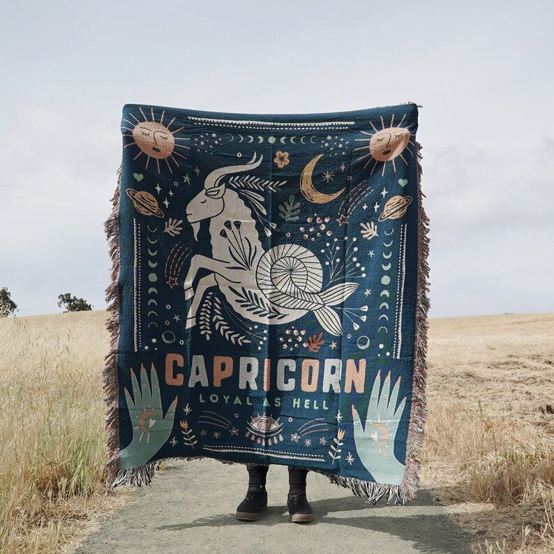 May include: Dark teal throw blanket featuring a Capricorn zodiac design. The blanket displays a goat illustration, sun, moon, and stars. Text reads "CAPRICORN LOYAL AS HELL". The blanket has a fringed edge.