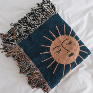 May include: A woven throw blanket with a blue background and a large orange sun with a face in the center. The sun has a closed eye and a smiling mouth. The blanket has a fringe border.