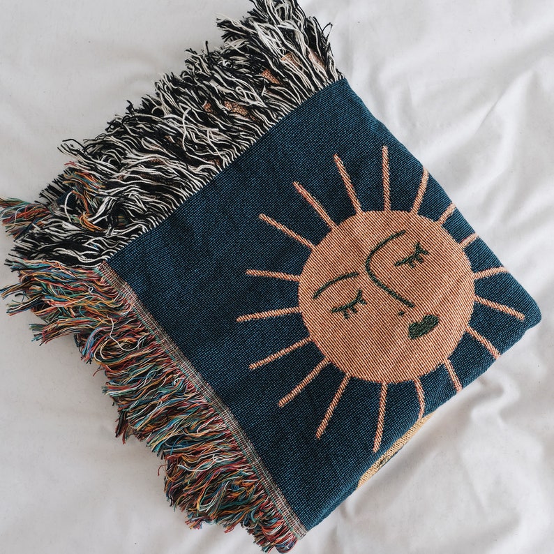 May include: A woven throw blanket with a blue background and a large orange sun with a face in the center. The sun has a closed eye and a smiling mouth. The blanket has a fringe border.