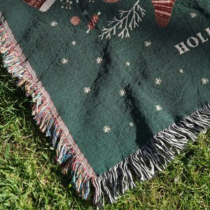 May include: Green woven throw blanket with a fringe border. The blanket features a white snowflake pattern and the word "Home" in white lettering.