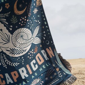 May include: A navy blue throw blanket with a Capricorn design. The blanket features a white seashell, celestial illustrations, and the words "CAPRICORN ROYAL AS HELL" in peach and white. The blanket has a fringed edge.