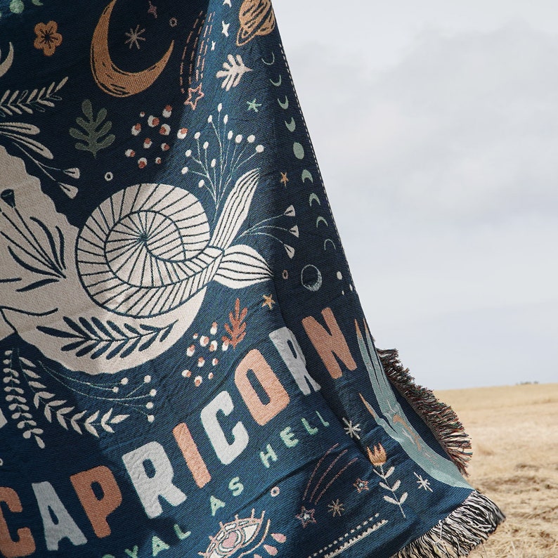 May include: A navy blue throw blanket with a Capricorn design. The blanket features a white seashell, celestial illustrations, and the words "CAPRICORN ROYAL AS HELL" in peach and white. The blanket has a fringed edge.