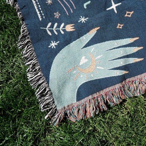 May include: A blue woven throw blanket with a hand design featuring a crescent moon and sun. The blanket has a fringe border.