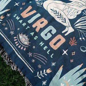 May include: A navy blue woven tapestry with the word "Virgo" in pink and blue lettering. The tapestry also features the text "Smart as Hell" and a stylized image of a person with a star and other celestial symbols.