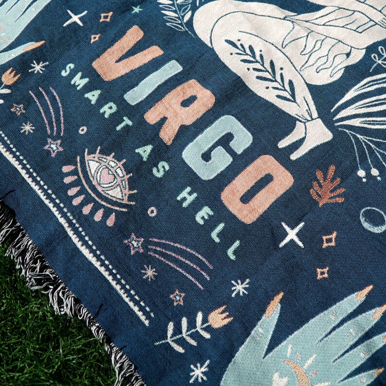 May include: A navy blue woven tapestry with the word "Virgo" in pink and blue lettering. The tapestry also features the text "Smart as Hell" and a stylized image of a person with a star and other celestial symbols.