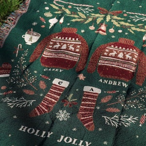 May include: Green throw blanket with a festive holiday design. The blanket features two red and white sweaters with the names "Casey" and "Andrew" embroidered on them. The blanket also features two red and white striped stockings with the letters "C" and "A" embroidered on them. The blanket has the words "Holly Jolly" embroidered on it.