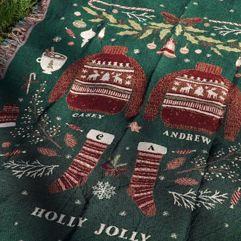 May include: Green throw blanket with a festive holiday design. The blanket features two red and white sweaters with the names "Casey" and "Andrew" embroidered on them. The blanket also features two red and white striped stockings with the letters "C" and "A" embroidered on them. The blanket has the words "Holly Jolly" embroidered on it.