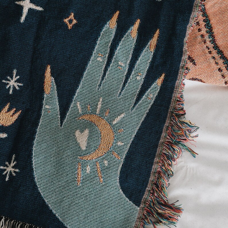 May include: A woven throw blanket featuring a teal hand design with gold fingernails, a crescent moon, and a sunburst. The blanket has a dark blue background with fringe along the edge. The design includes stars and other celestial elements.