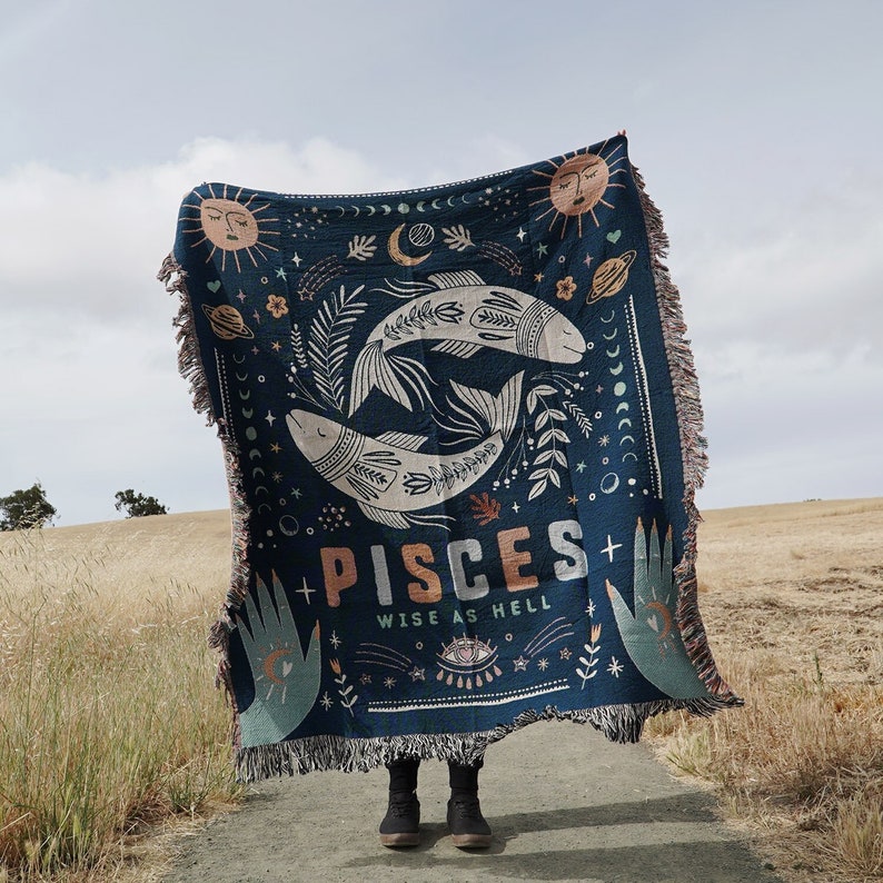 May include: A navy blue woven throw blanket featuring a Pisces zodiac design. The blanket displays two fish, suns, moons, and the text "PISCES WISE AS HELL". The blanket has a fringed edge and is held up against a natural outdoor backdrop.