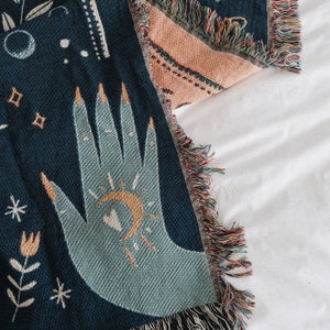 May include: A blue woven throw blanket with a hand and moon design. The hand is light blue and the moon is yellow. The blanket has a fringe border.