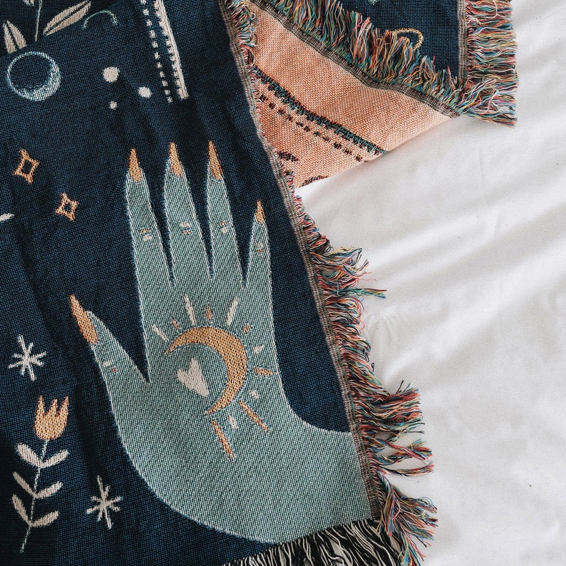 May include: A blue woven throw blanket with a hand and moon design. The hand is light blue and the moon is yellow. The blanket has a fringe border.